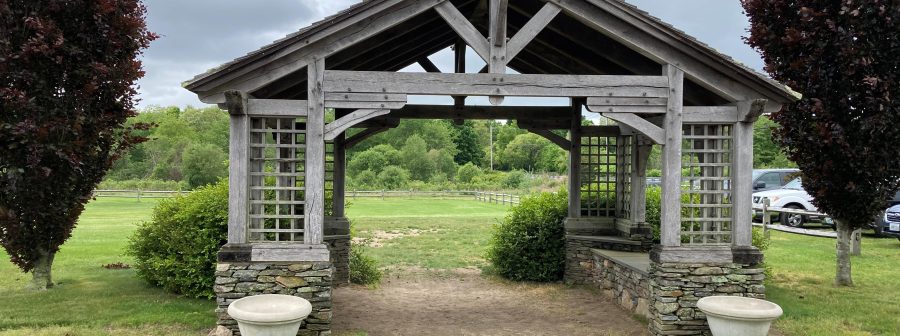 The front of a weathered timber frame pavilion featuring embellished beams, stone foundation and benches.