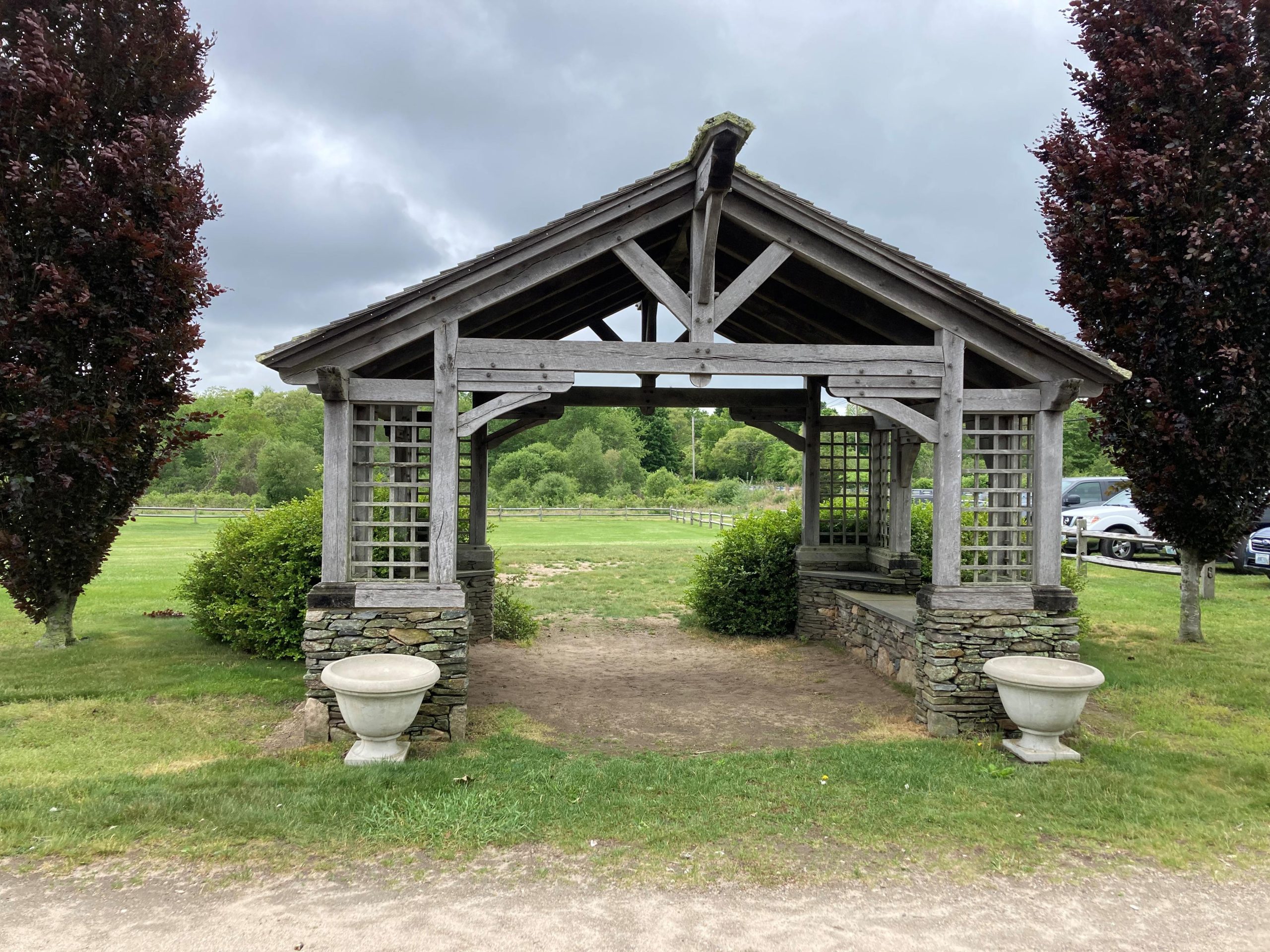 The front of a weathered timber frame pavilion featuring embellished beams, stone foundation and benches.