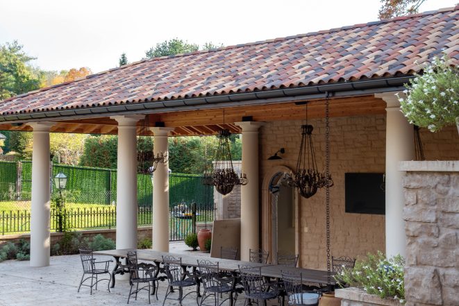 A custom timber framed pavilion with a clay tile roof, outdoor seating, custom lighting, and stone pillars in a Mediterranean style