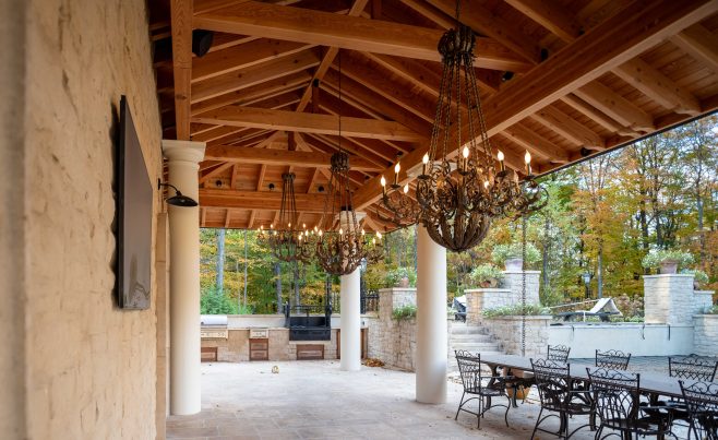 A custom timber framed pavilion with a clay tile roof, outdoor seating, custom lighting, and stone pillars in a Mediterranean style