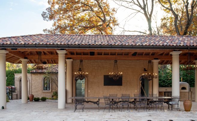 A custom timber framed pavilion with a clay tile roof, outdoor seating, custom lighting, and stone pillars in a Mediterranean style