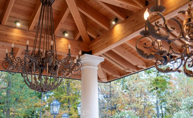 A custom timber framed pavilion with a clay tile roof, outdoor seating, custom lighting, and stone pillars in a Mediterranean style