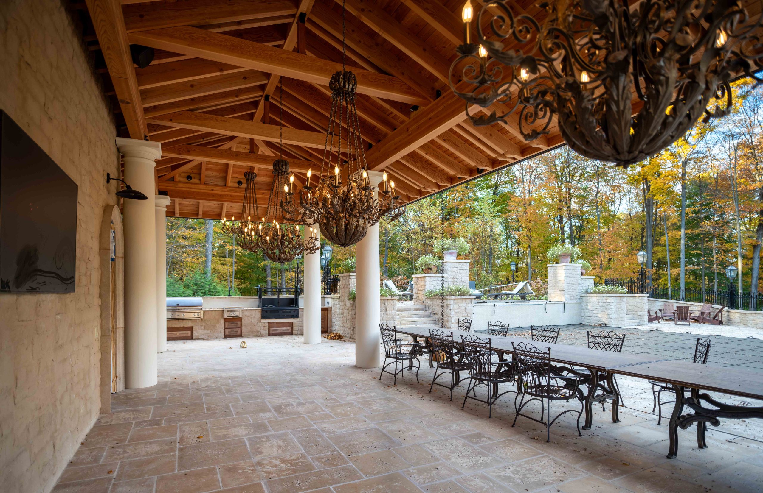 A custom timber framed pavilion with a clay tile roof, outdoor seating, custom lighting, and stone pillars in a Mediterranean style
