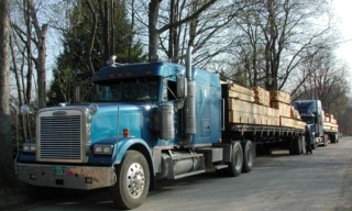 Blue semi-truck hauling a lumber trailer on a tree-lined street; another truck follows behind.